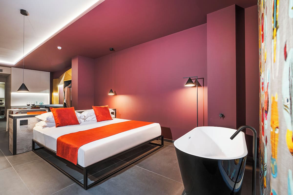 A modern hotel room with a bed, bedside tables, a bathroom with a sink, and a view of the kitchen area.