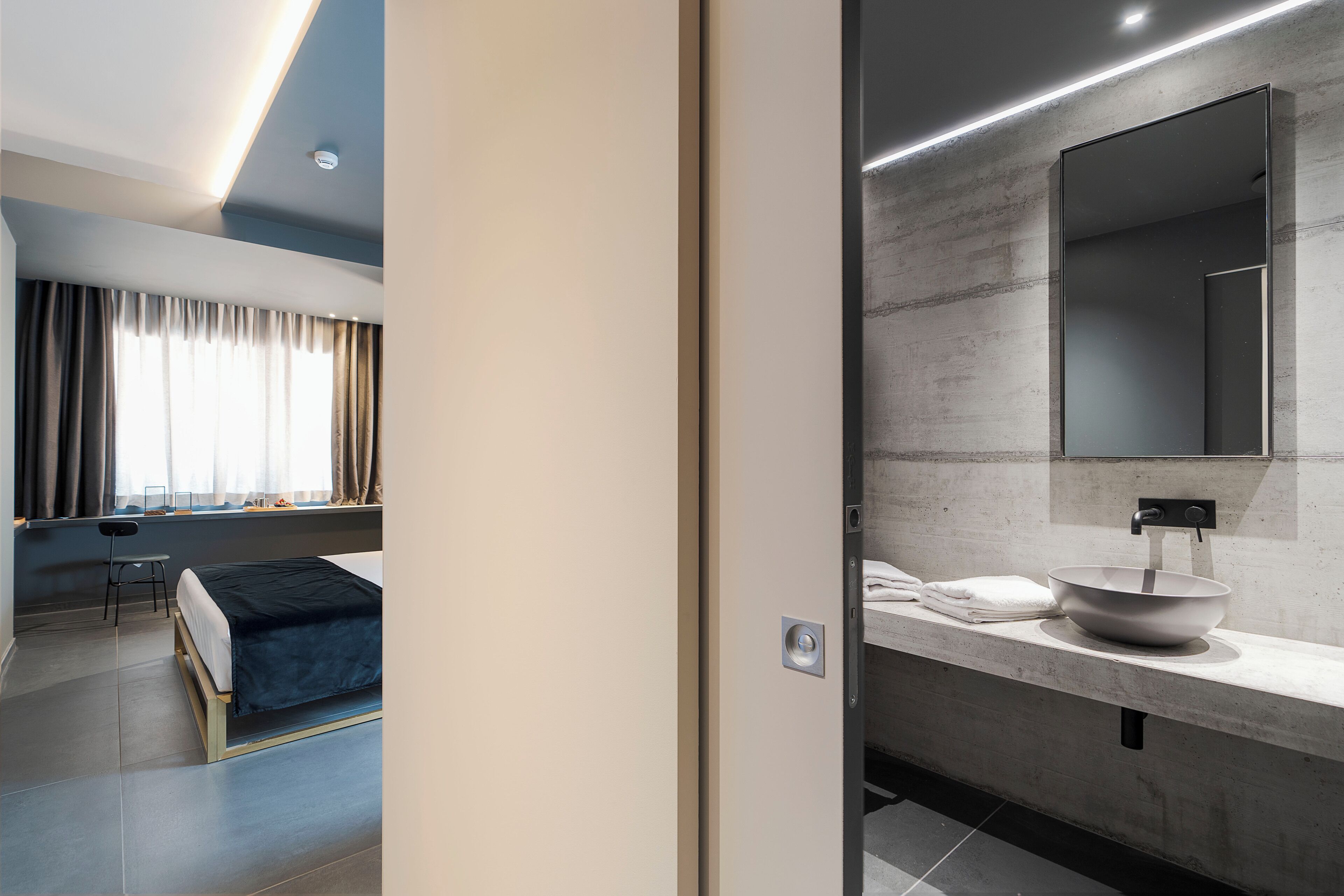 A modern hotel room with a bed, a desk, and a bathroom with a large mirror and sink.