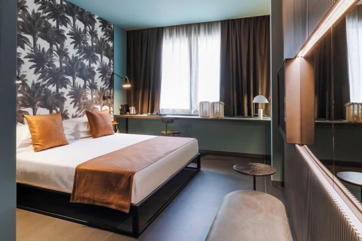 Una habitación de hotel moderna con una cama grande, una pequeña mesa redonda, un banco, una ventana con cortinas y una pared con papel tapiz tropical.