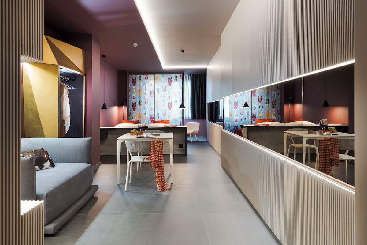A modern hotel room with a sofa, a dining area, a mirror, and a wardrobe.