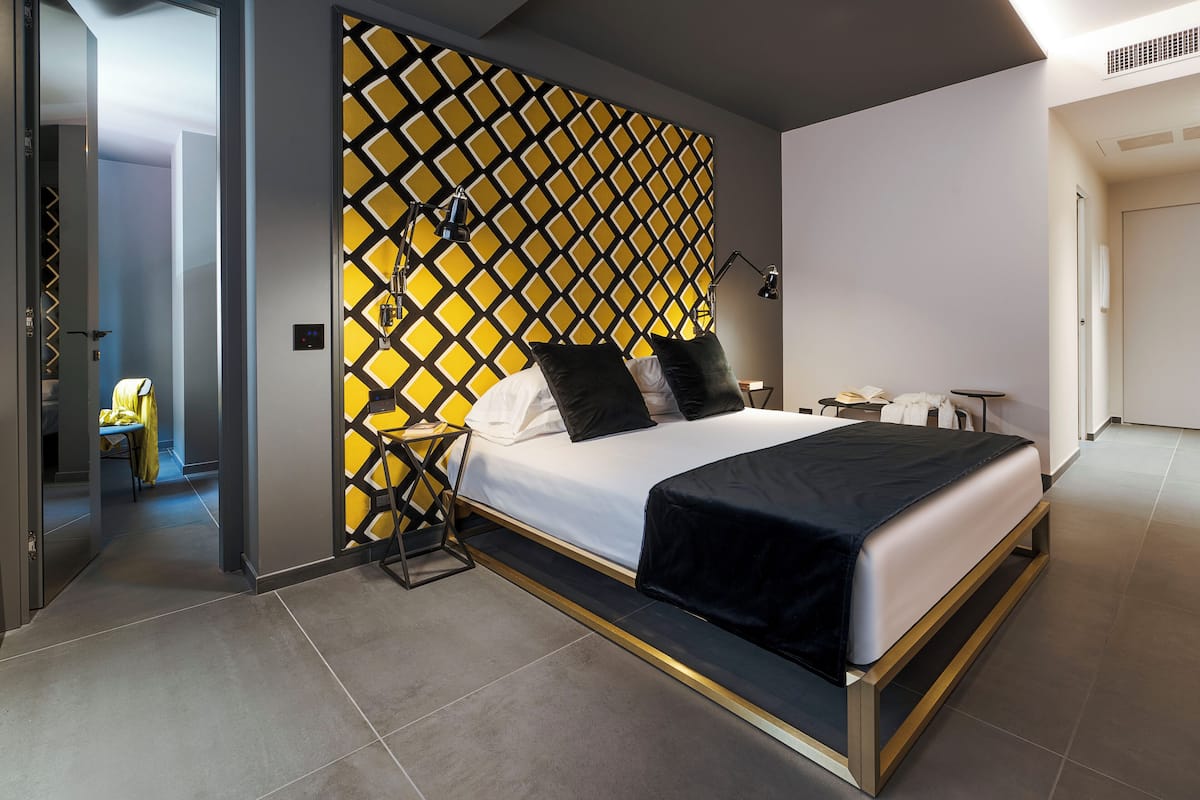 A modern hotel room with a large bed, a bedside table, a lamp, and a decorative wall panel.