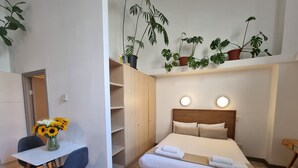 A bedroom with a bed, bedside tables, a desk with a chair, a vase of sunflowers, and potted plants on a shelf. - Mutual Heights M15 by CTHA (Cape Town)