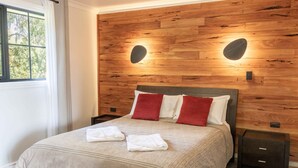 A bedroom with a wooden headboard, two bedside tables, a bed with a beige bedspread, two red pillows, and two folded towels. - Scarlet Woods Chalets (Quinninup)