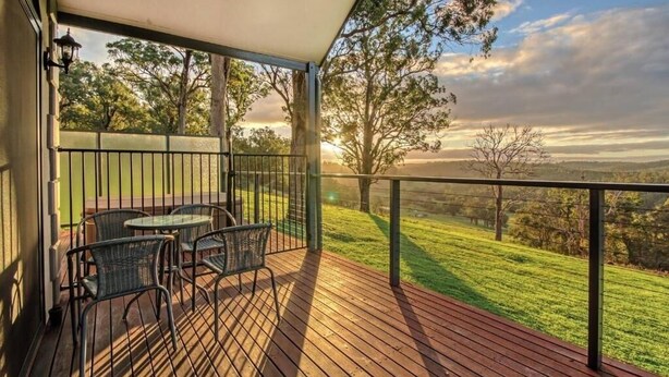 A wooden deck with outdoor furniture, a railing, and a view of a grassy landscape with trees. - Scarlet Woods Chalets (Quinninup)