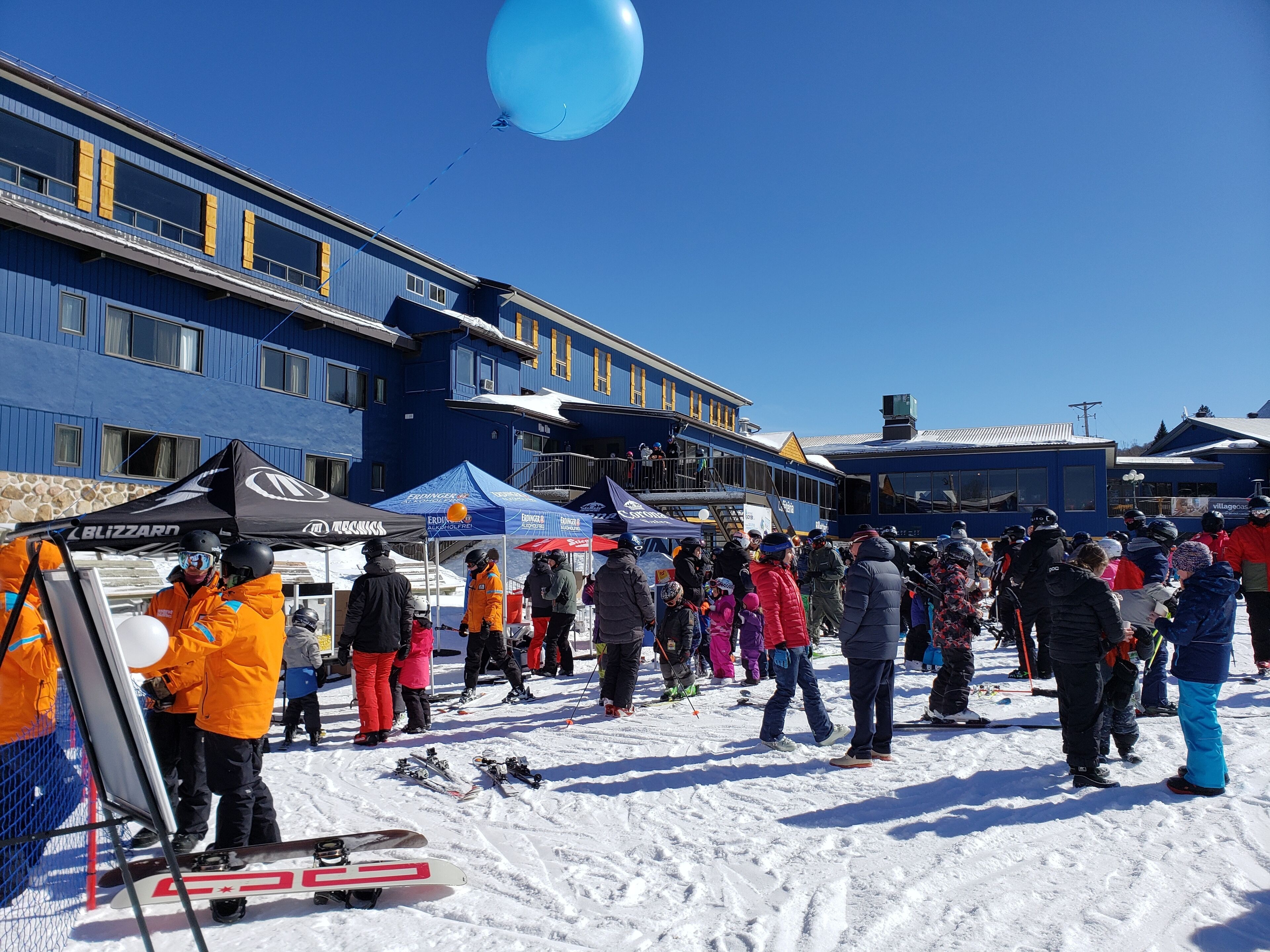 A snowy outdoor area with a crowd, tents, and a building with blue walls and yellow windows.