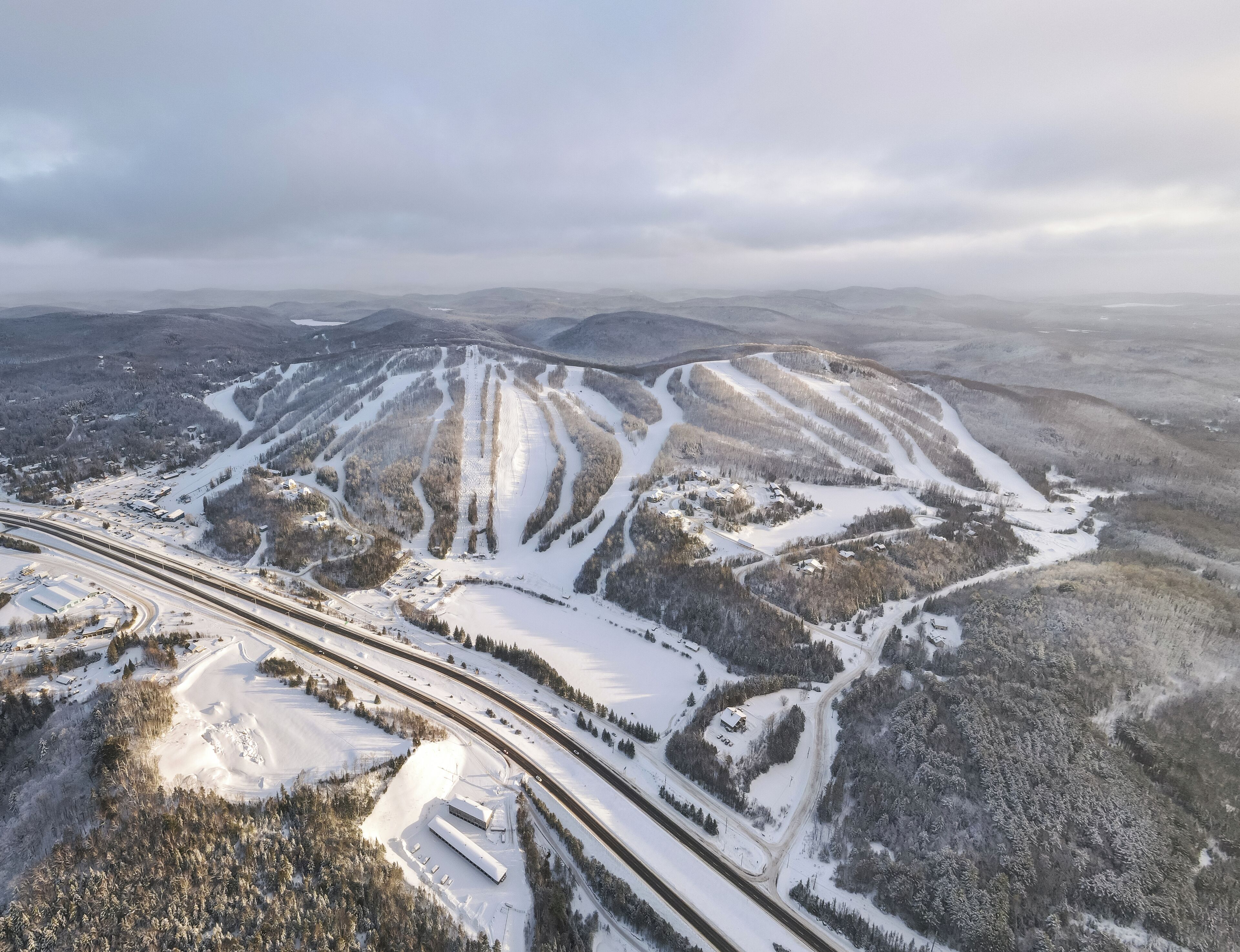An aerial view of a snow-covered ski resort with ski runs, buildings, and a highway.