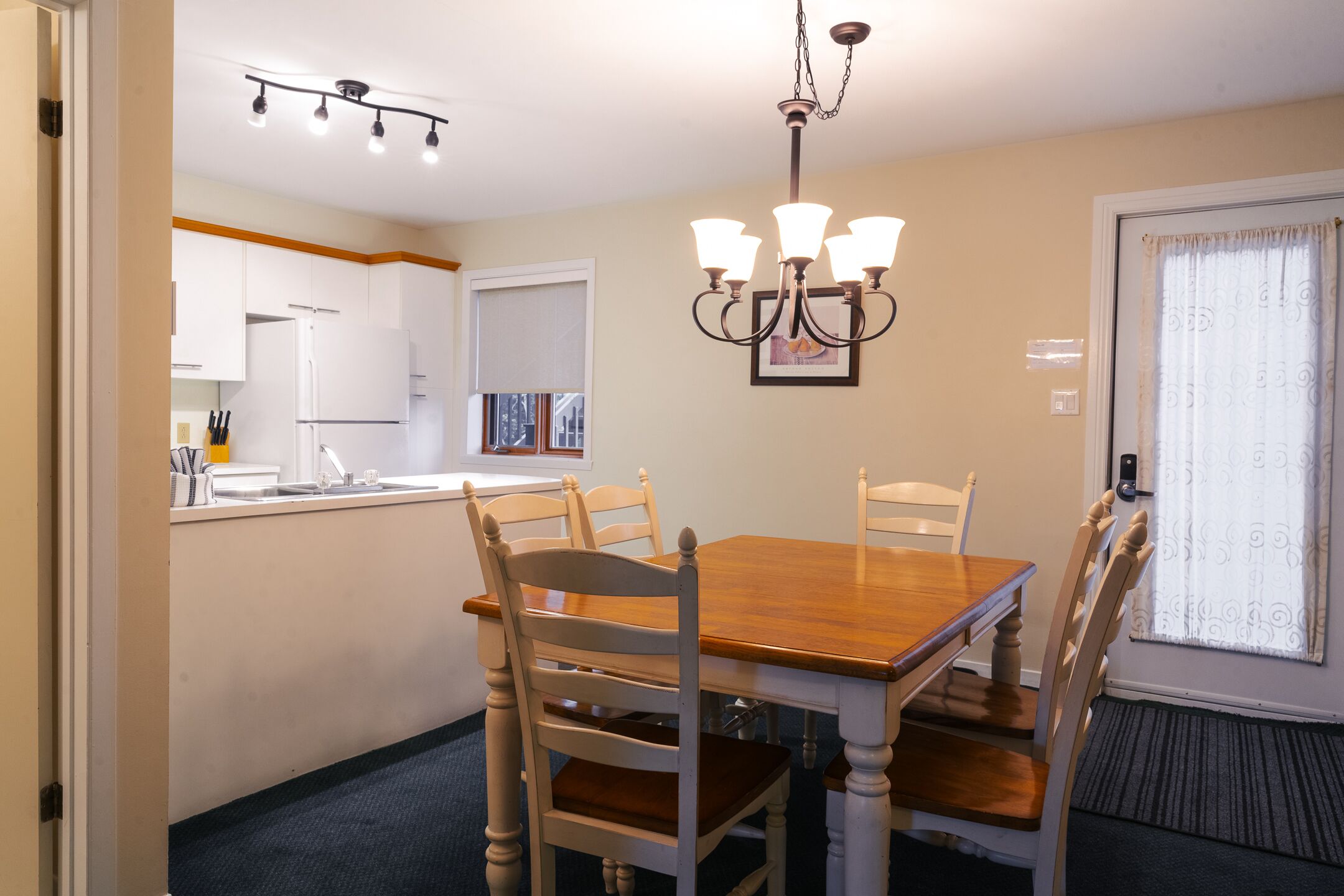 A dining area with a wooden table and white chairs, a chandelier, and a kitchen in the background.