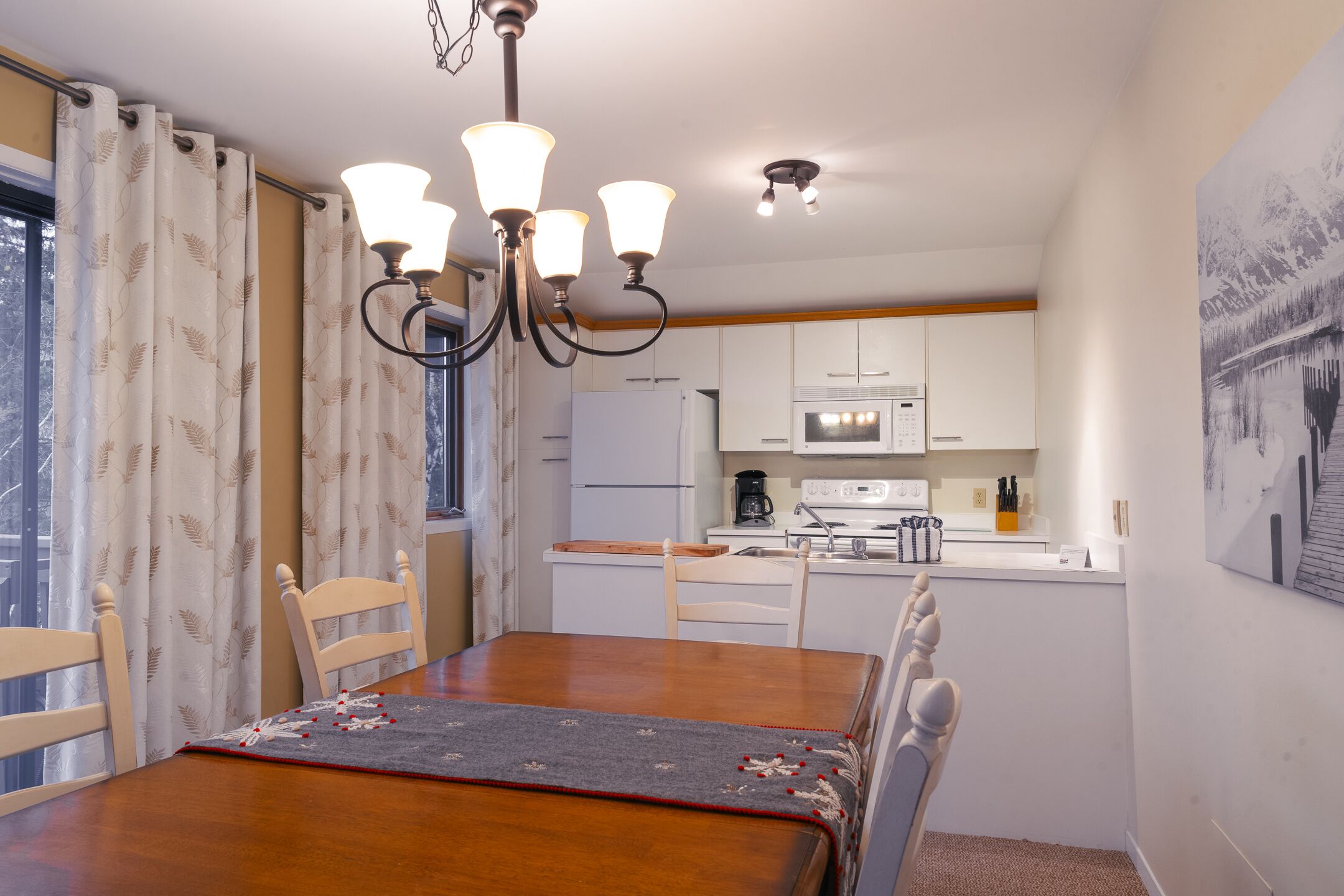 A dining area with a wooden table, chairs, and a chandelier. There is a kitchen in the background with white cabinets and appliances.