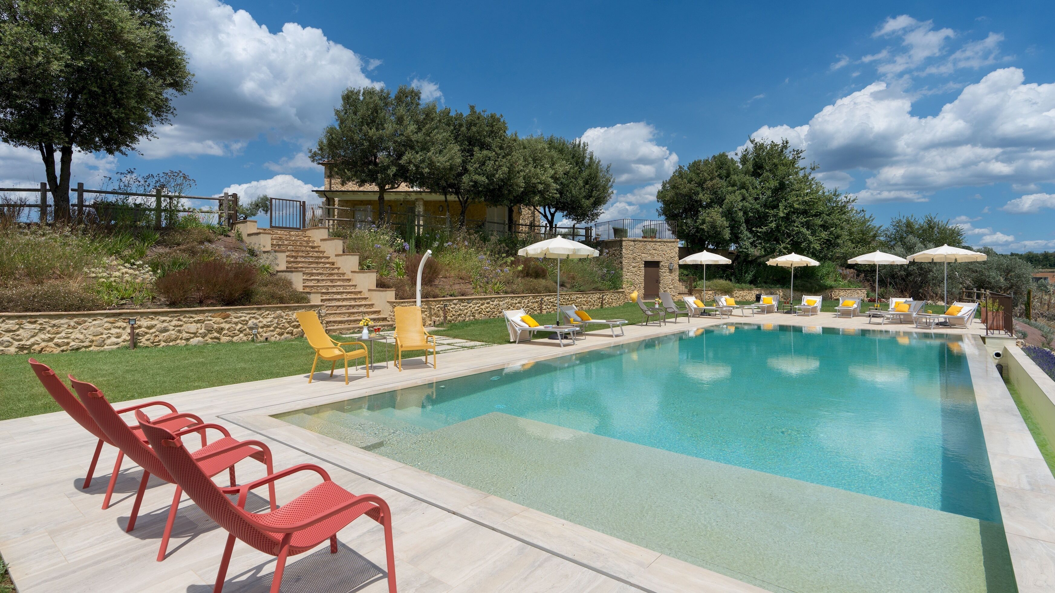 Piscina con tumbonas y sombrillas, rodeada de un muro de piedra y vegetación.