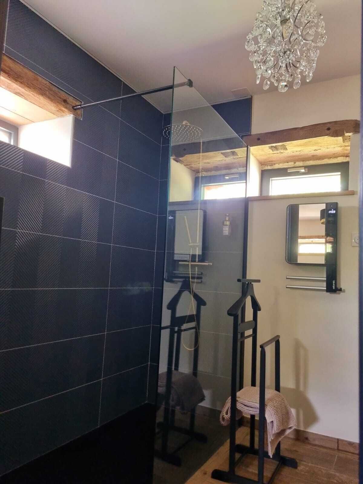 A modern bathroom with dark tiled walls, a glass shower enclosure, a wooden ceiling, and a chandelier.