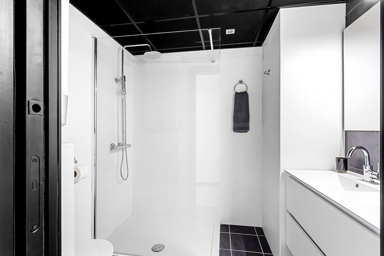 A modern bathroom with a glass-enclosed shower, a white sink with a mirror, and a black tiled floor.