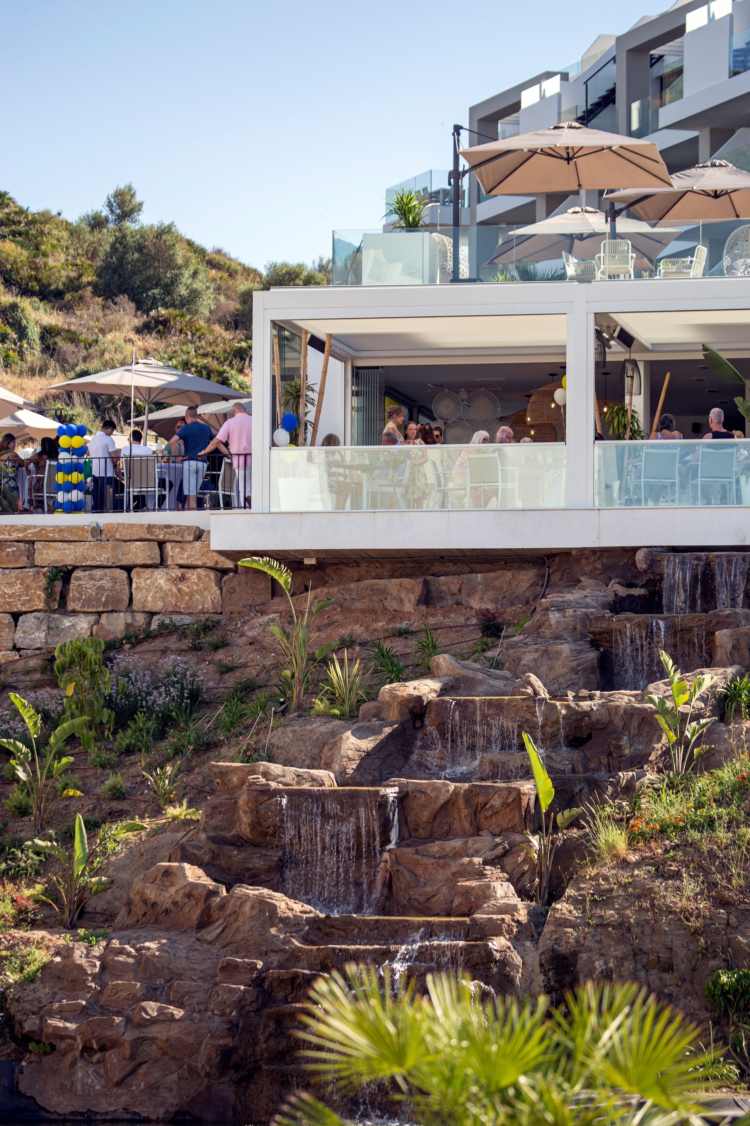 A modern outdoor dining area with a waterfall feature, stone walls, and a glass railing overlooking the terrace.