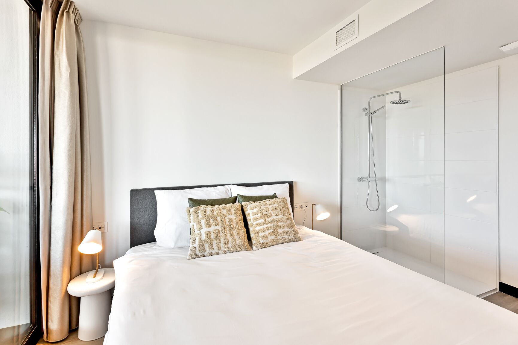 Una habitación de hotel moderna con una cama grande, cabina de ducha de vidrio y una mesita de noche con lámpara.