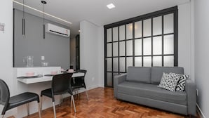 A modern living room with a grey sofa, black chairs, a wooden herringbone floor, a white desk with a computer, and a large window with a black frame. - Apto 2 quartos no Centro de BH (Belo Horizonte)