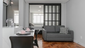 A modern living room with a grey sofa, a dining table with pink bowls, and a large window. - Apto 2 quartos no Centro de BH (Belo Horizonte)