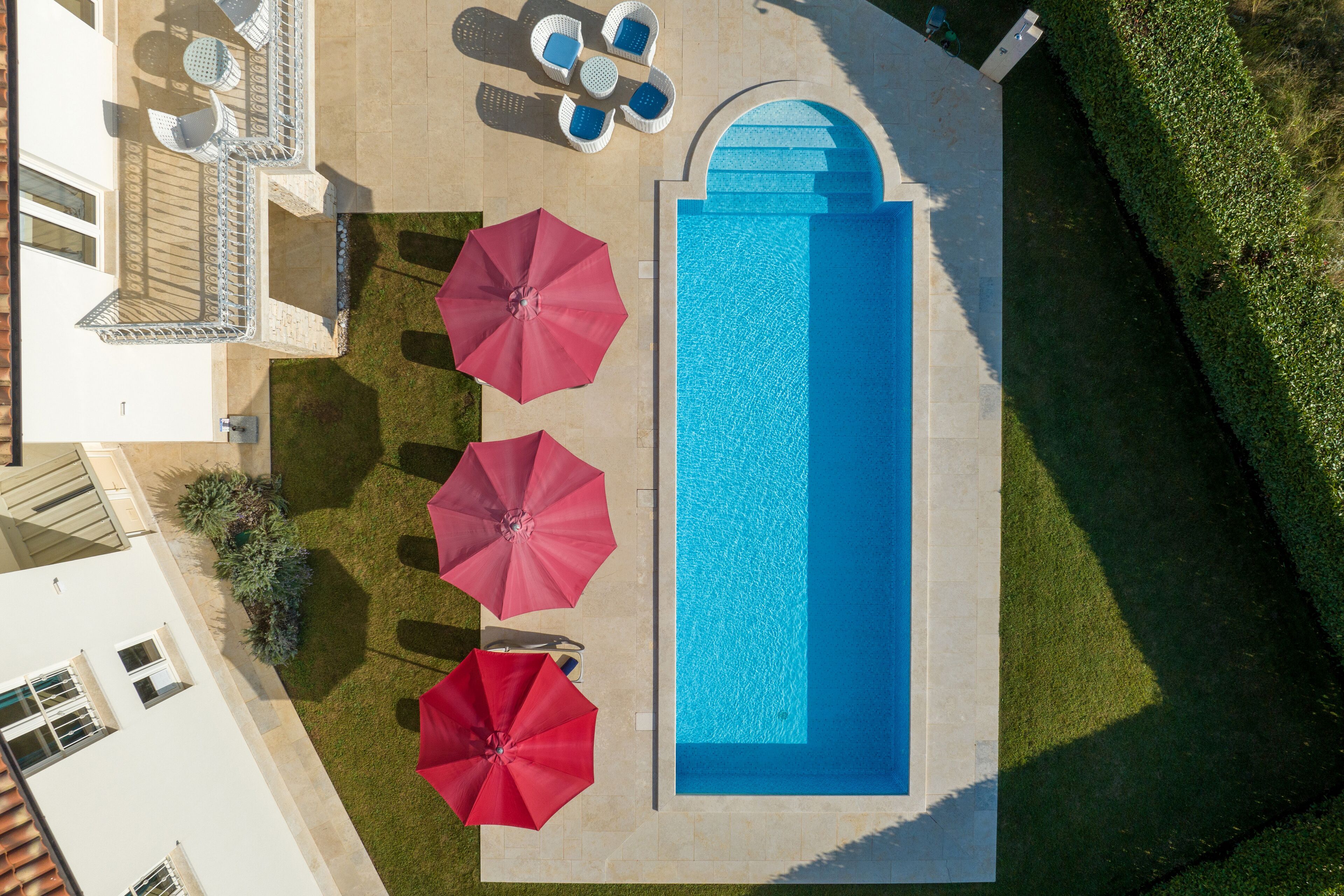 An aerial view of a swimming pool area with a sun lounger, two red umbrellas, and a seating area with white chairs.