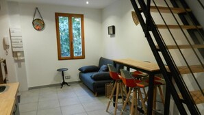 A modern living room with a sofa, a small round table, and two colorful chairs. A wooden staircase is visible to the right. - The little house (Ceyreste)