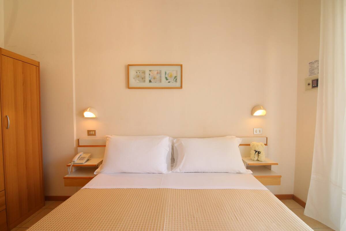 A hotel room with a large bed, two bedside tables, a wooden wardrobe, and a framed picture on the wall.