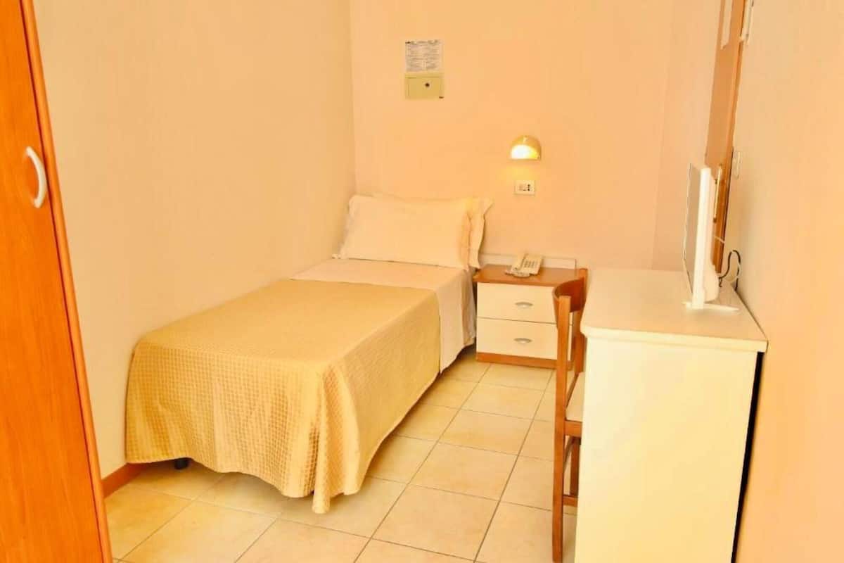 A small, neatly arranged bedroom with a bed, a bedside table with a lamp, a small cabinet, and a mirror.