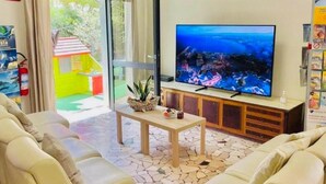 A living room with a large flat-screen TV, beige sofas, a coffee table, and a view of a cityscape through a glass door. - Hotel Soave Miriam (Rimini)