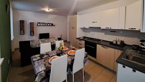 Una cocina compacta con armarios blancos, una mesa de comedor para cuatro personas y una pequeña sala de estar con un sofá y televisor.