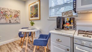 A compact kitchen with a gas stove, a coffee bar sign, a framed painting, and a dining area with blue chairs and a small table. - 'the Dragonfly House' - Walk to Downtown Pensacola (Pensacola)