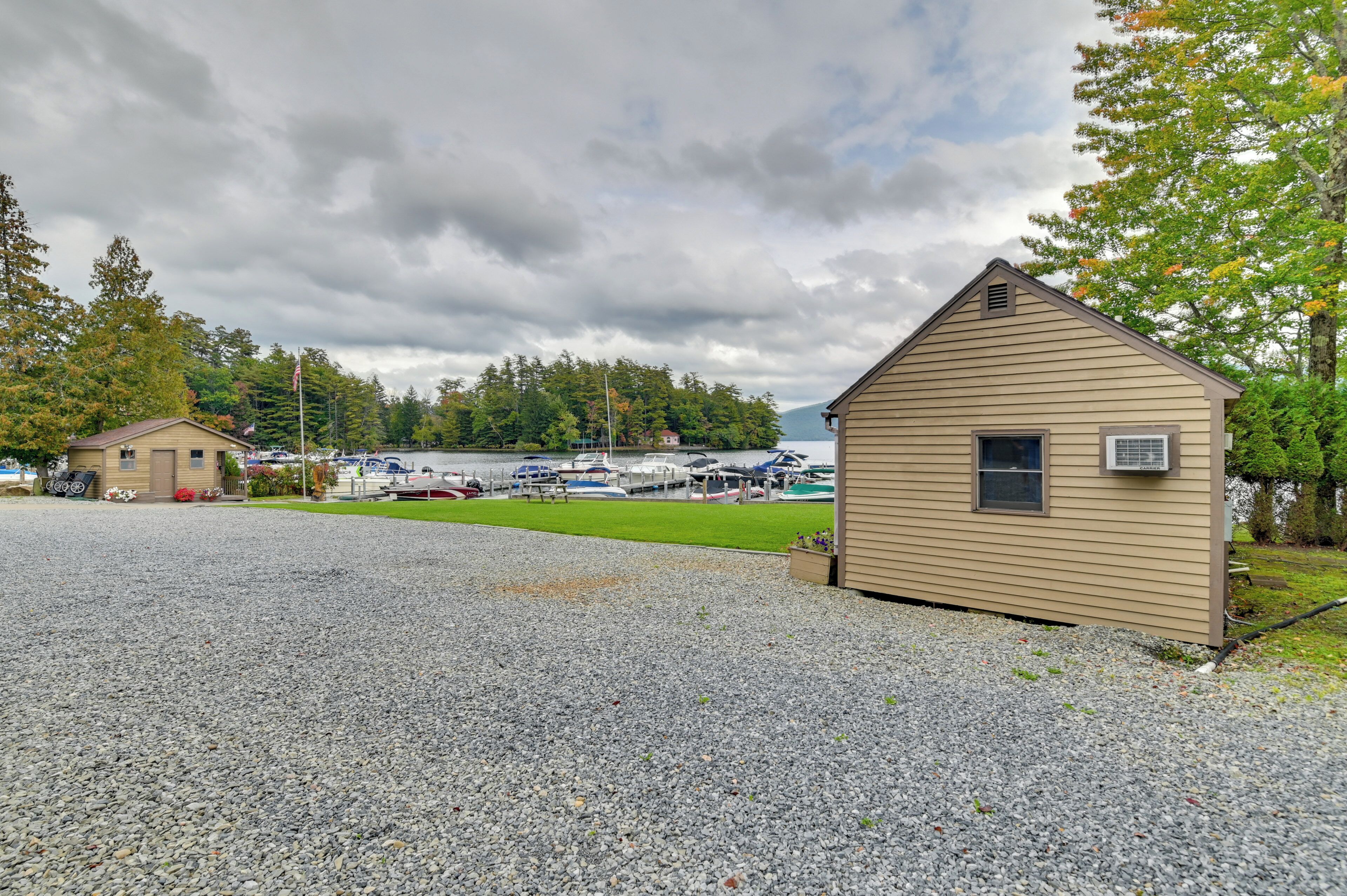 A gravel parking area with a small wooden building, boats docked in the background, and a lake.