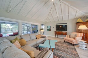 A spacious living room with a large sofa, a coffee table, and a TV. There are large windows and a ceiling with exposed beams.