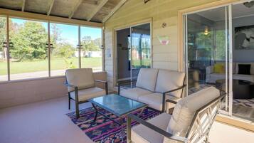 Relax in this spacious living area with a sofa set, coffee table, and patterned rug, surrounded by large windows offering views of the surrounding greenery.