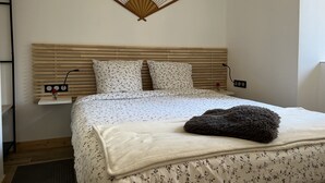 A neatly made bed with a floral bedspread, a bedside table with a vase of flowers, and a wall-mounted fan. - Appartement Avec Sauna et Wifi, au Coeur du Village, Avec vue sur la Montagne (Le Bourg-d'Oisans)