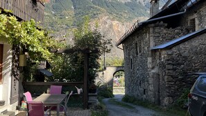 A rustic outdoor seating area with a stone building, a wooden pergola, and a mountain backdrop. - Appartement Avec Sauna et Wifi, au Coeur du Village, Avec vue sur la Montagne (Le Bourg-d'Oisans)