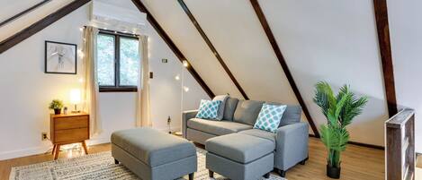 Un dormitorio estilo loft acogedor con techo inclinado, un sofá seccional gris, un reposapiés a juego, un tapete estampado, una mesita de noche con lámpara y una planta en maceta.