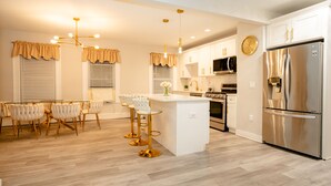 Une cuisine moderne avec des meubles blancs, un réfrigérateur en acier inoxydable, un îlot central doté de tabourets, et un coin repas avec une table et des chaises en verre.