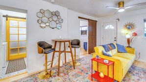Una sala de estar con un sofá amarillo, una mesita auxiliar roja, una mesa redonda con dos sillas y una alfombra colorida.