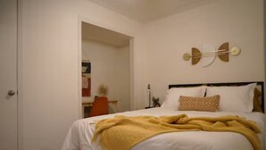A bedroom with a bed, a bedside table, a wall-mounted light fixture, and a door leading to another room. - Sedona Retreat at La Hacienda (Wichita Falls)
