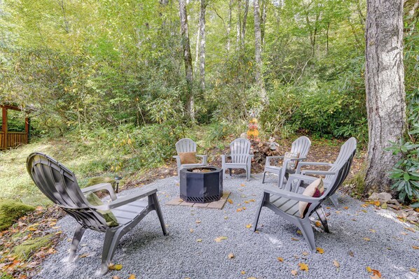 A circular fire pit surrounded by six Adirondack chairs with cushions, set on a gravel surface in a forested area.