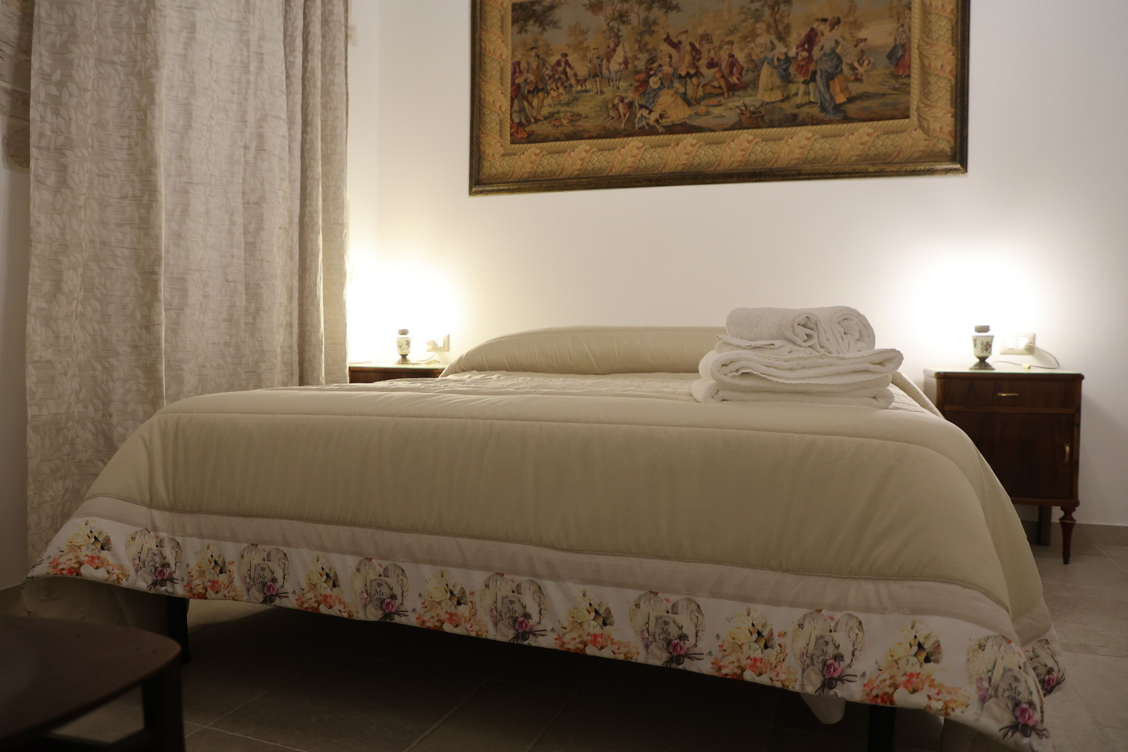 Un lit bien fait, recouvert d’une couvre-lit à motifs floraux, un tableau accroché au mur et une table de chevet avec une lampe.