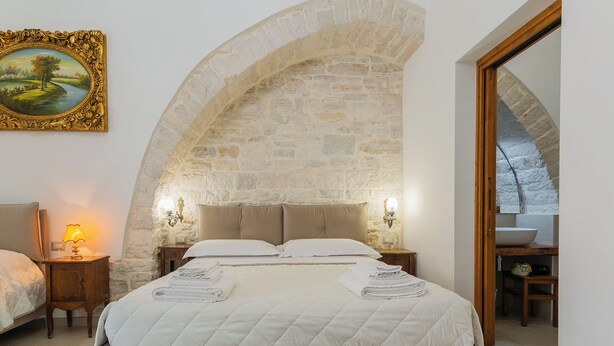 A bedroom with a stone arch, a bed with white bedding, two bedside tables with lamps, and a painting on the wall. - Antico Monastero (Bitonto)