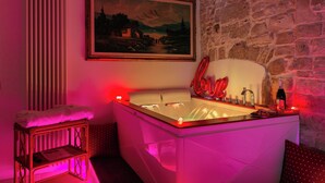 A room with a stone wall, a painting, a bathtub with a red 'Love' sign, a bottle, and a small table with a towel. - Antico Monastero (Bitonto)