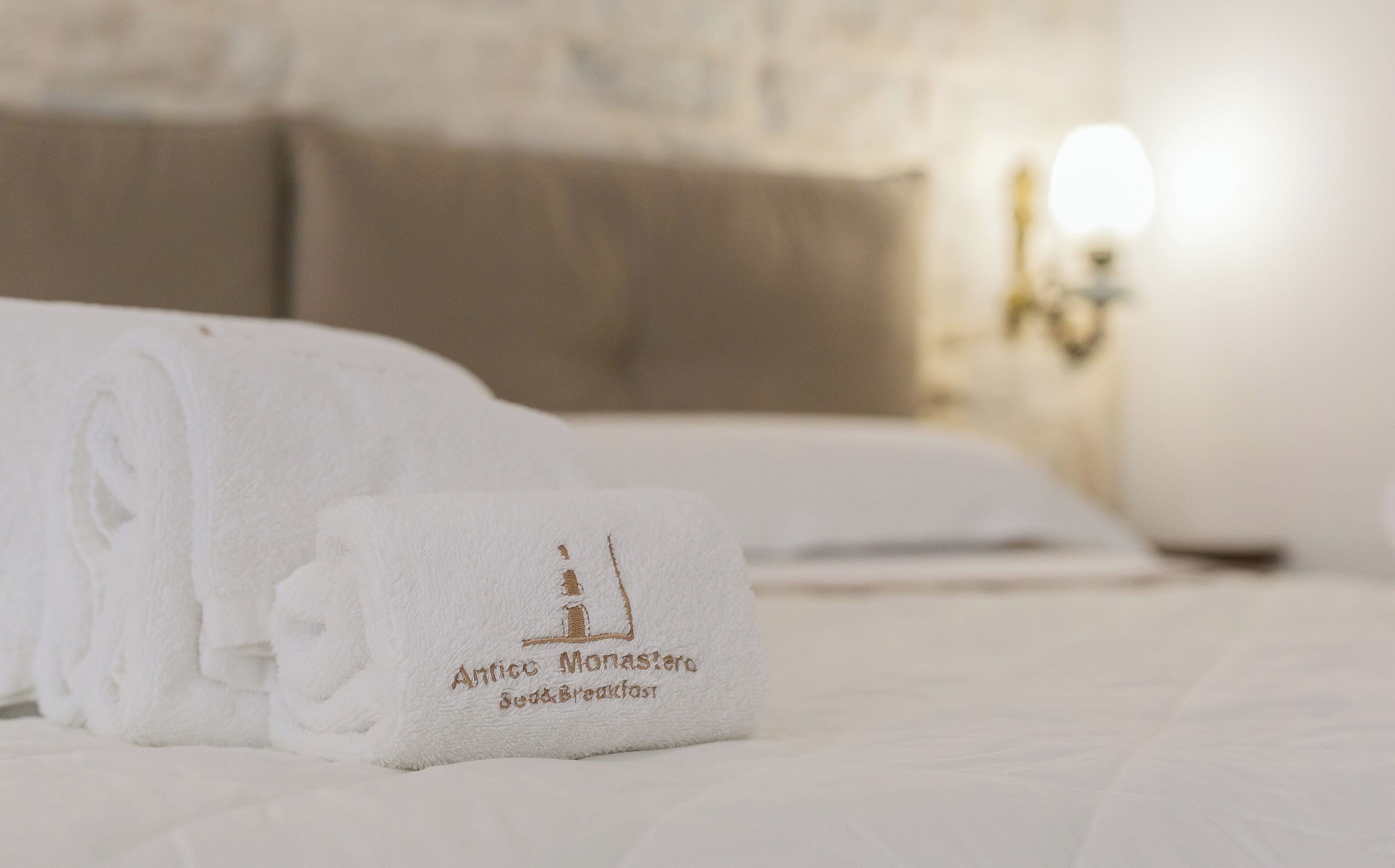 A neatly made bed with a folded towel and a smaller towel with the text "Antico Monastero Bed & Breakfast" on it.