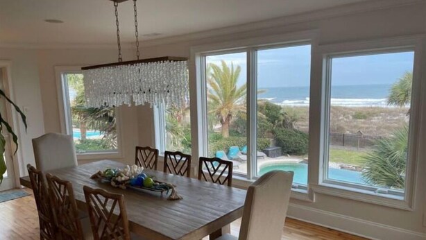 Un espace repas comprenant une table en bois, des chaises et un lustre. De grandes fenêtres offrent une vue sur l’océan et une piscine.