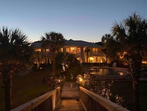 A large, two-story house with a swimming pool, surrounded by palm trees and a wooden deck leading to the entrance.