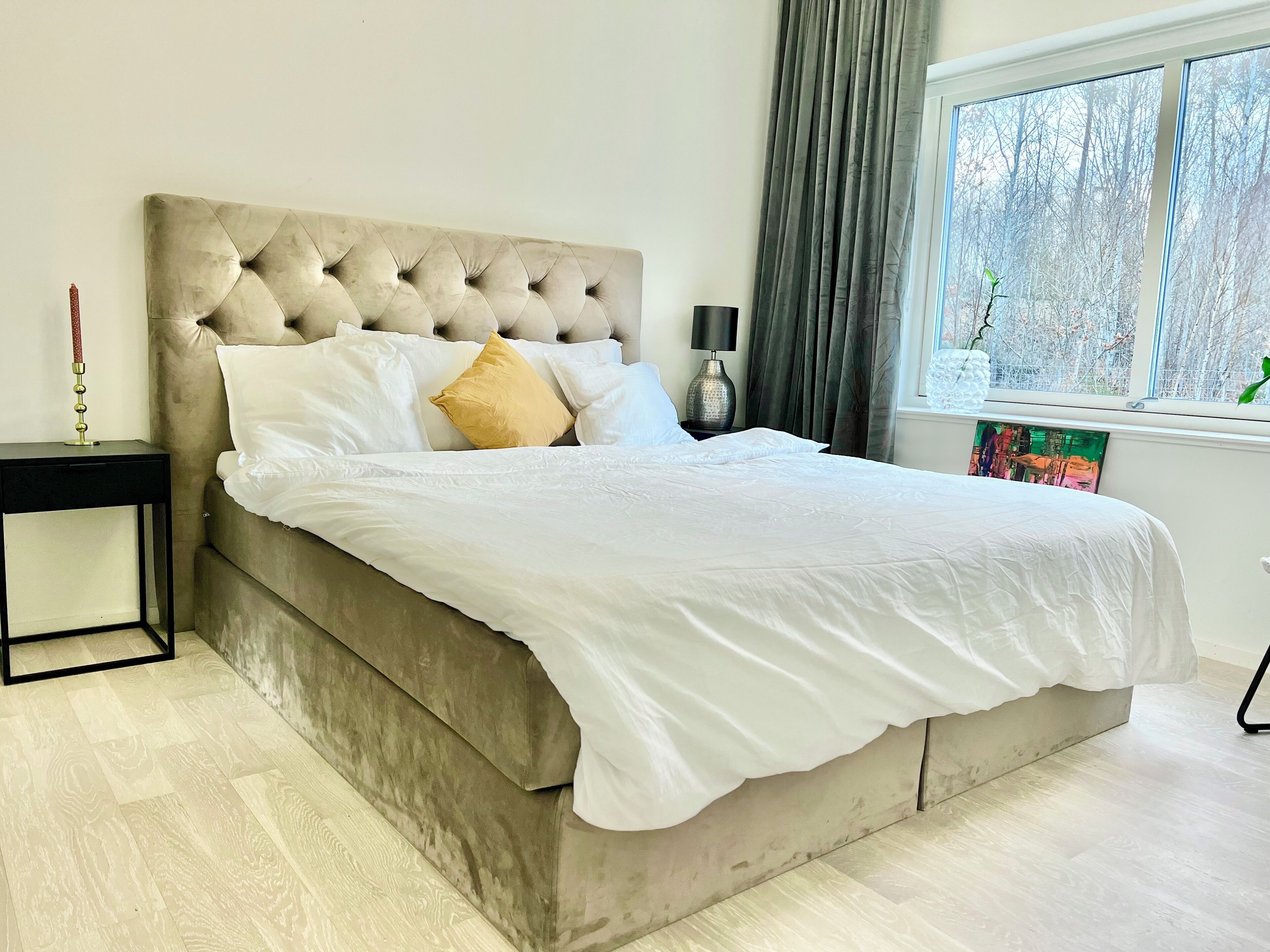 A bedroom with a tufted headboard, a bed with white bedding, a bedside table with a lamp, and a window with a view of trees.