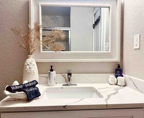 Ein Badezimmer mit weißem Waschbecken, einem Spiegel mit dekorativem Rahmen, einer Vase mit getrockneten Blumen, einer Handseifenflasche und einem Handtuch mit der Aufschrift 'Makeup Makeup'.