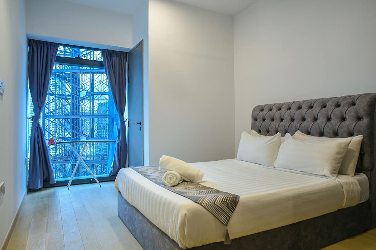 A neatly made bed with a grey headboard, a window with blue curtains, and a view of a building structure outside.