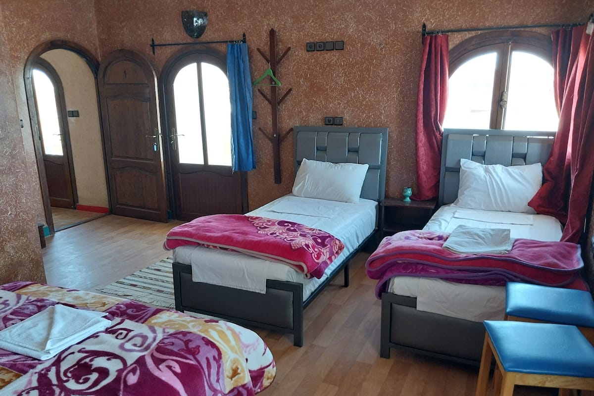 Quarto de hotel com duas camas de solteiro, piso de madeira, uma pequena mesa e uma cadeira. Há duas janelas com cortinas e uma porta com design em arco.