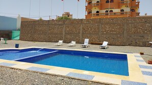 A swimming pool with lounge chairs, a stone wall, and a hotel building in the background. - Sud-Est Palace guest House Kalaa m'gouna (Ait Sedrate Sahl El Gharbia)