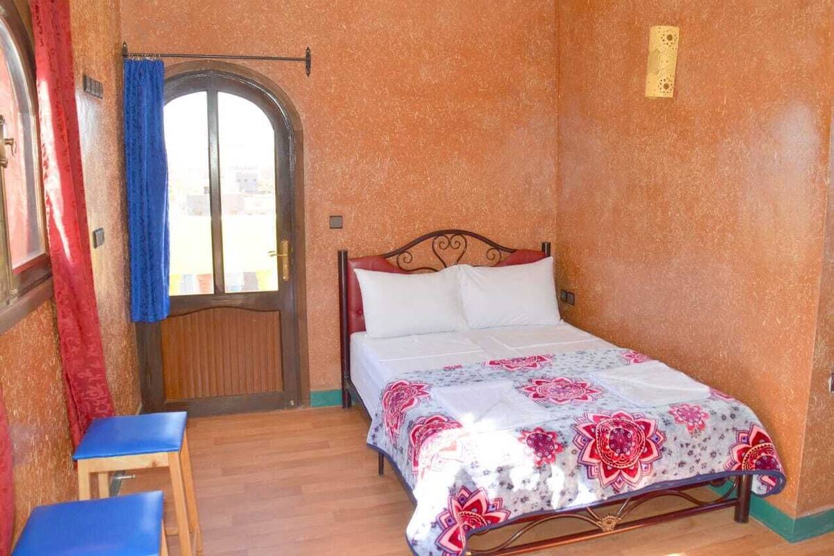 Um quarto com uma cama, uma cadeira de madeira, uma janela com cortinas e uma porta.