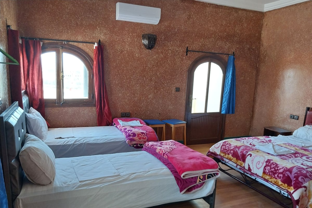 Quarto de hotel com duas camas, uma mesa pequena, uma cadeira e uma janela com cortinas.