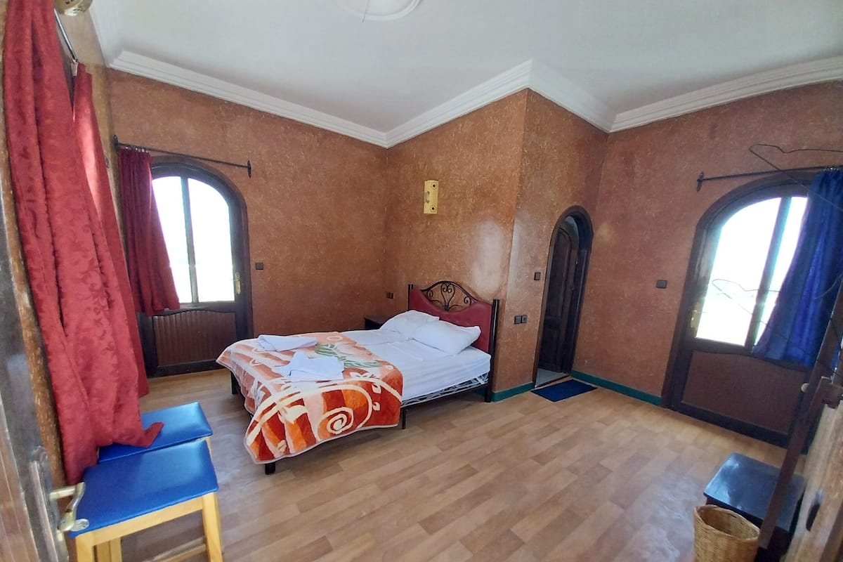 Quarto com uma cama, uma mesinha e uma cadeira. Há duas janelas com cortinas, uma de cada lado da cama.